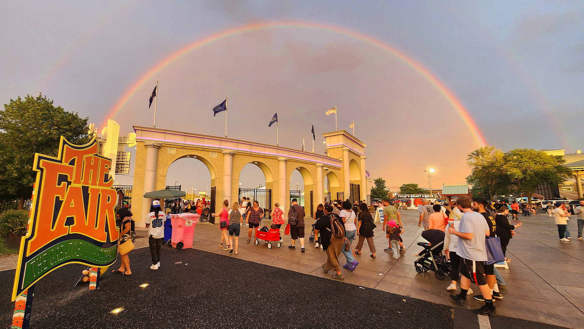 Fairgoers walking through main gates with a rainbow against a cloudy sky in the background.