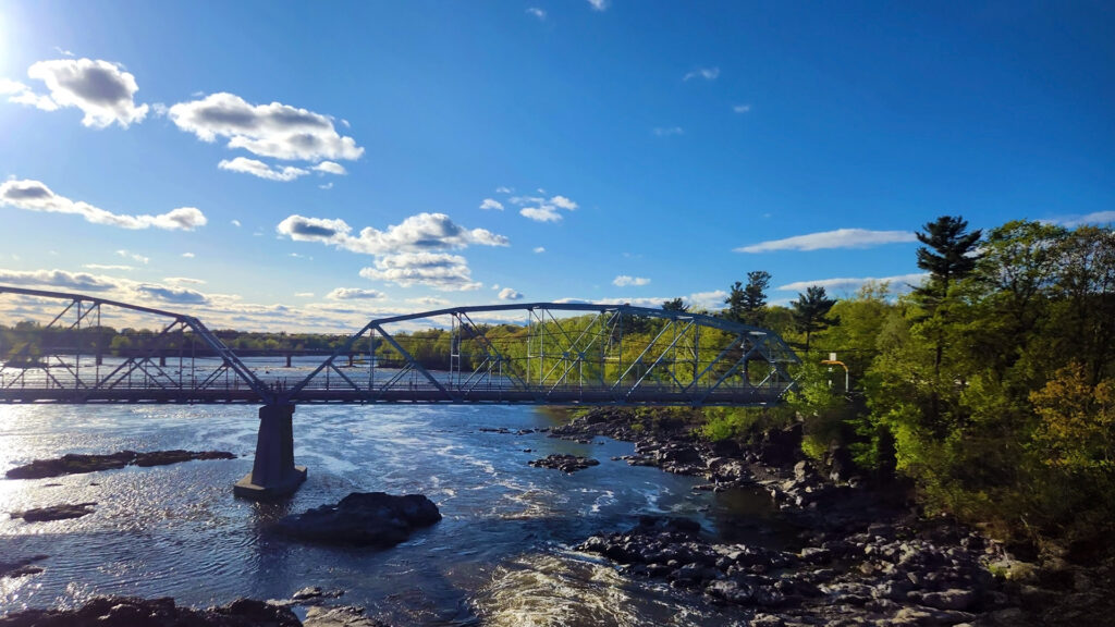 Crossing a river with a bridge in the foreground against blue skies and green trees on the river side.