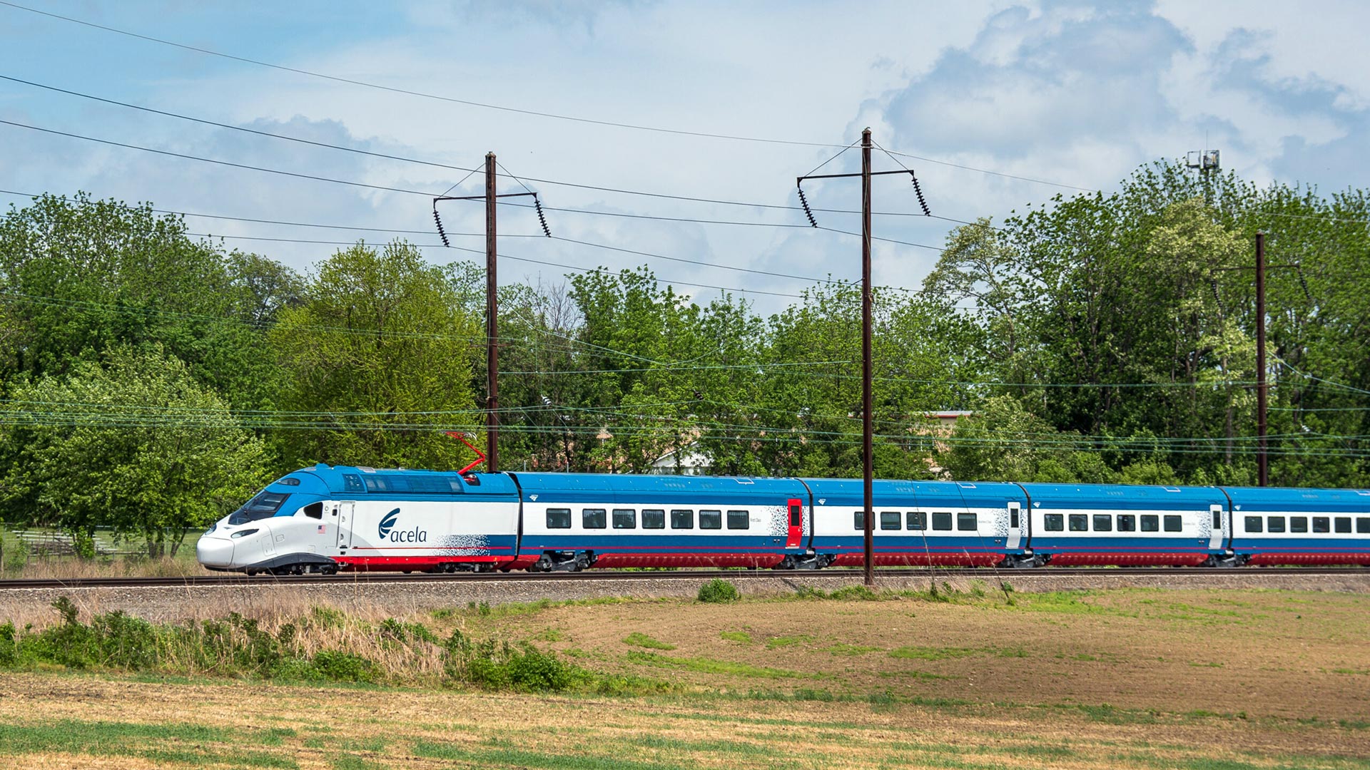 New white and blue Amtrak NextGen Acela rounding a curve through a field.