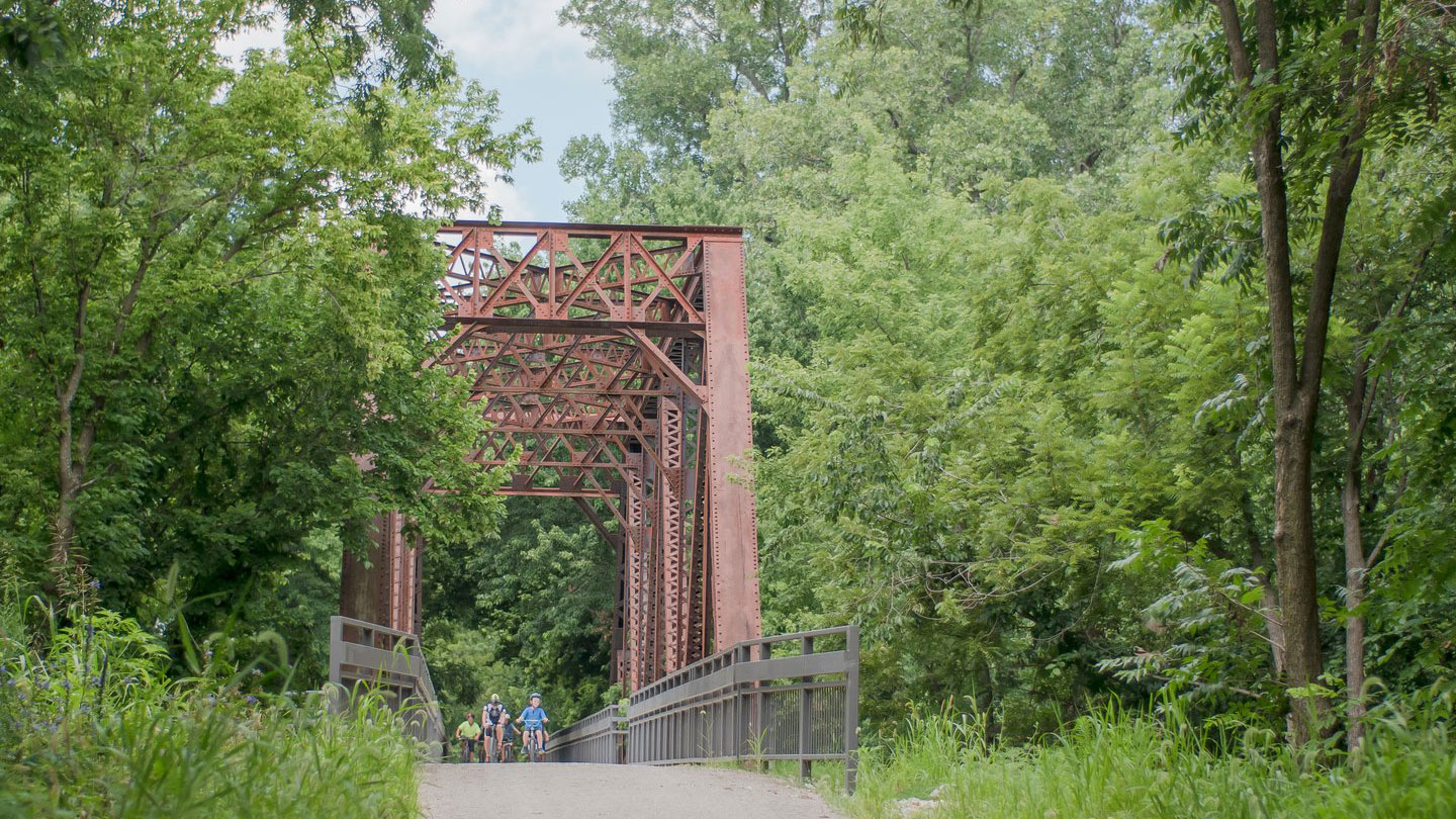 Bike riders crossing brown steel bridge with trees lining each side of the trail.