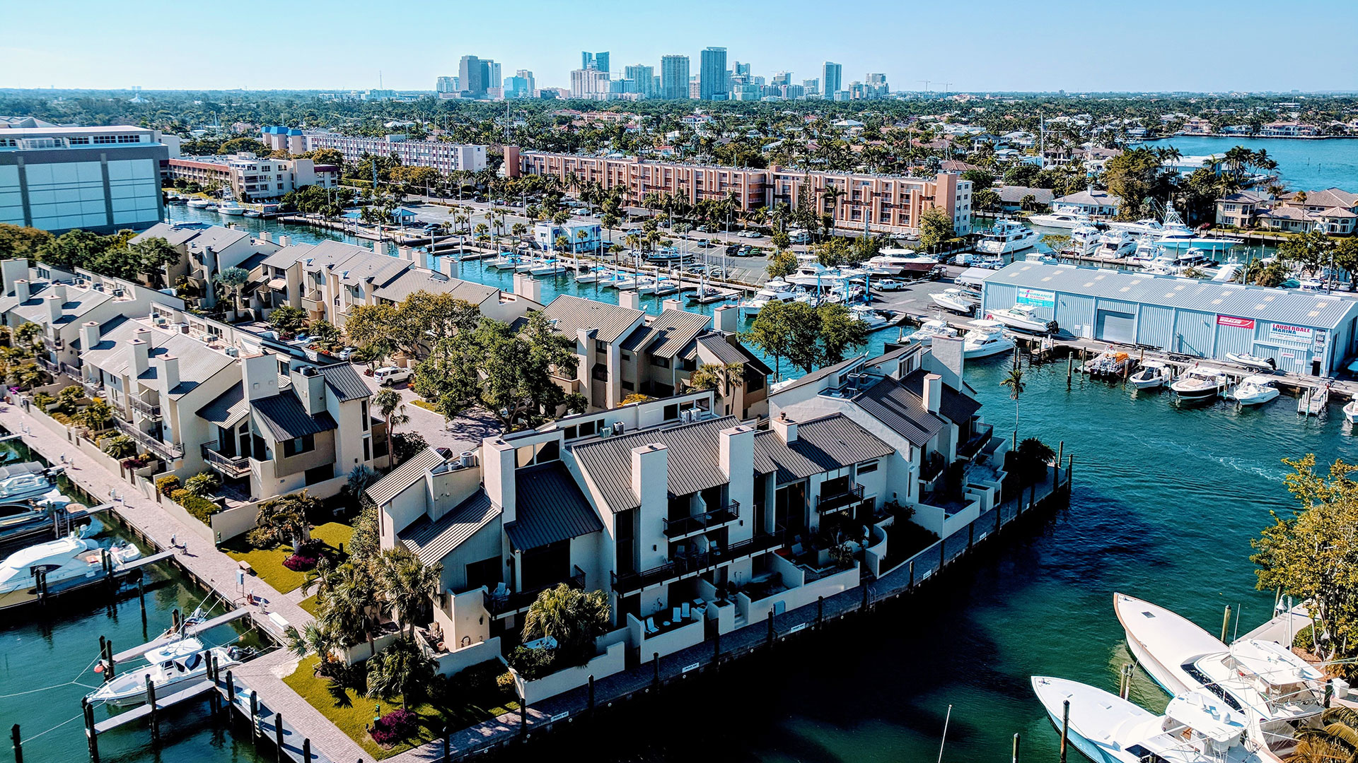 Houses along the shores of a harbor with boats in slips and downtown Fort Lauderdale in the background.