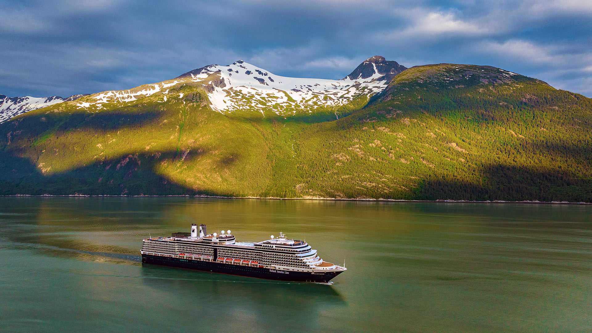 Cruise ship travels through a fjord with the sun shining on the green mountain in the background.