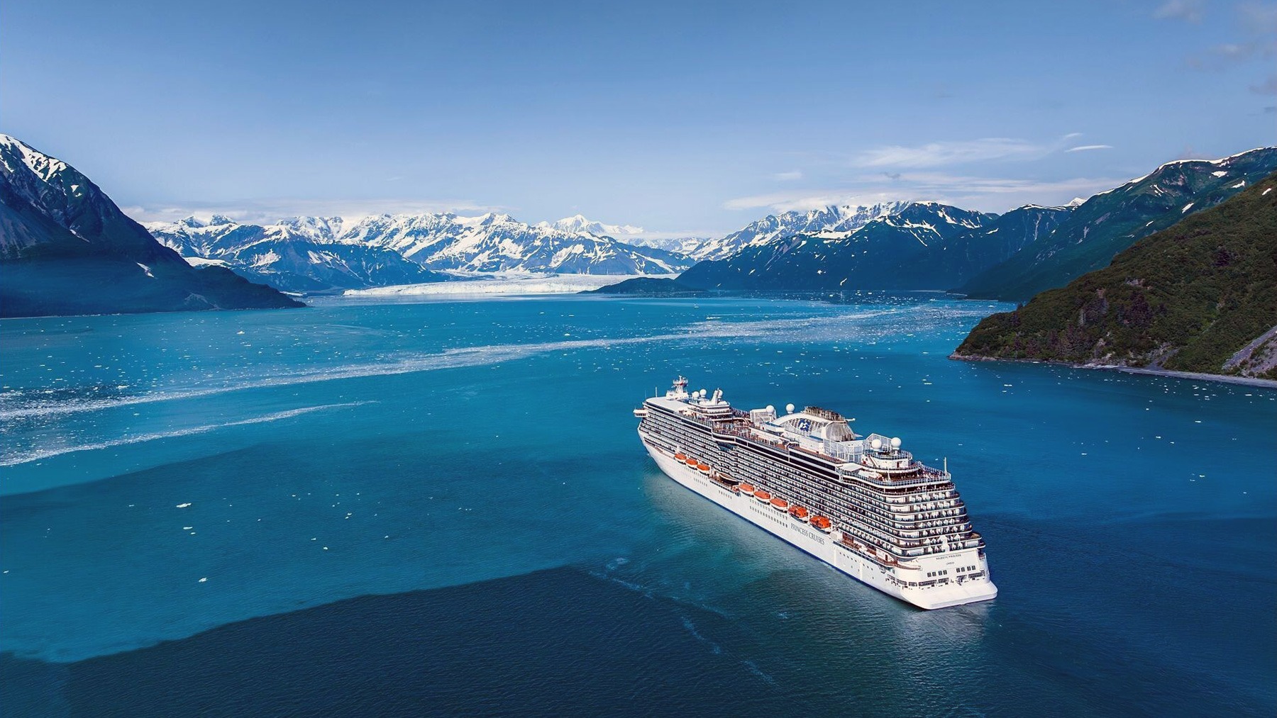 Cruise ship sailing in crystal blue waters in Alaska with glaciers in the background.