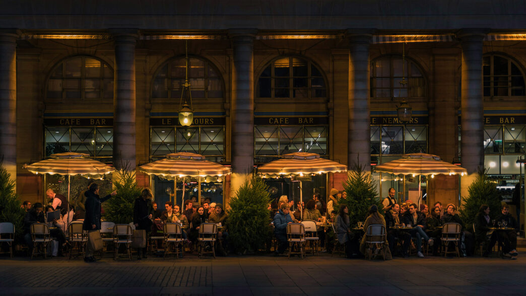 Individuals sitting at various tables at a restaurant outside under warmly lit umbrellas.