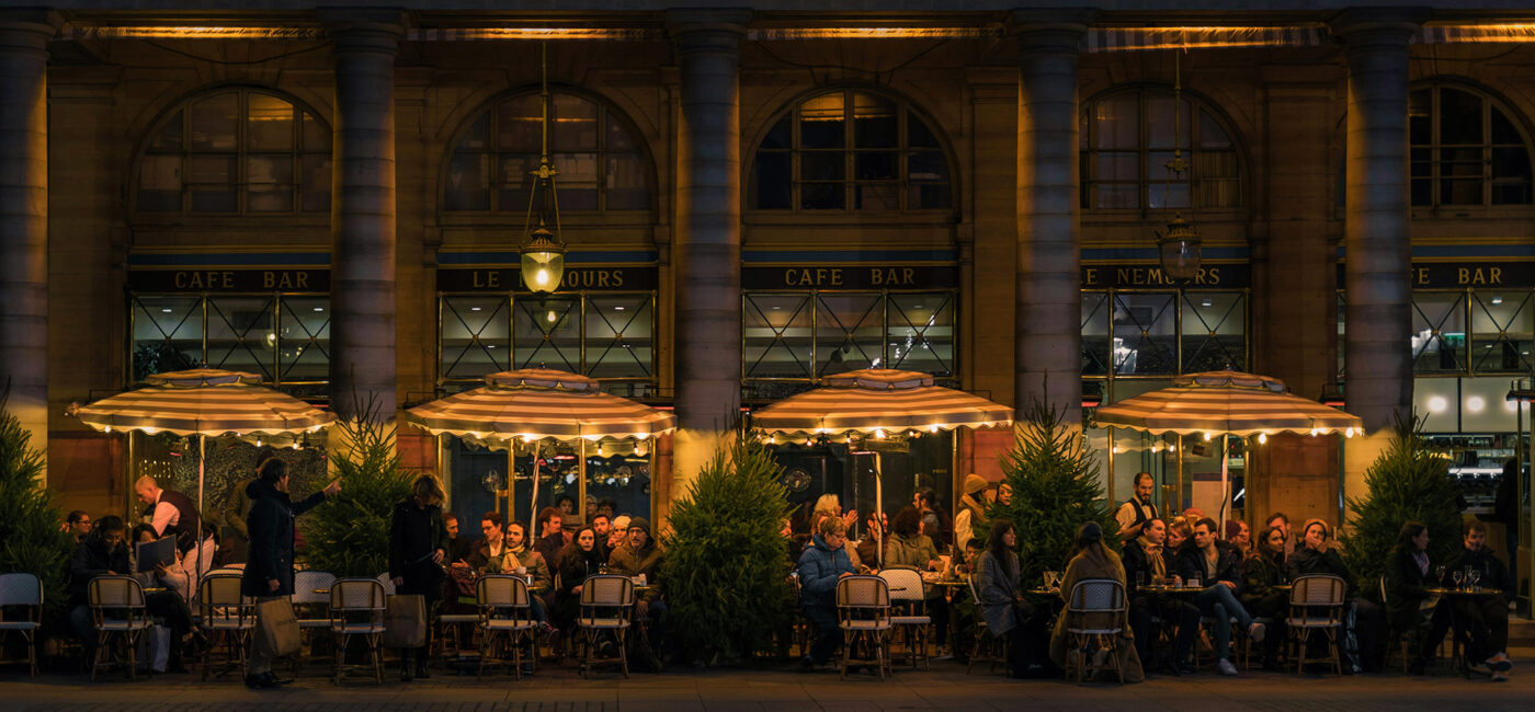 Individuals sitting at various tables at a restaurant outside under warmly lit umbrellas.