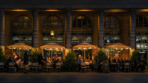 Individuals sitting at various tables at a restaurant outside under warmly lit umbrellas.