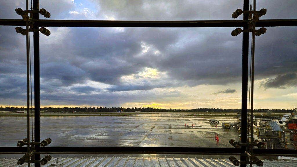 Looking out through floor to ceiling windows at Sea-Tac airport watching the sunset with grey clouds and sun in the distance.