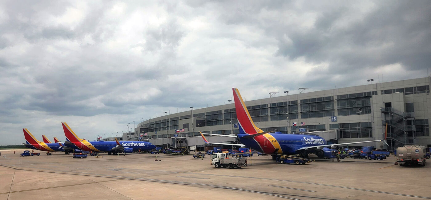 Several Southwest airplanes are parked at a jet bridge/ground equipment on a wide airport tarmac under a cloudy sky, with a modern terminal building in the background.