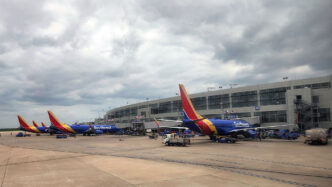 Several Southwest airplanes are parked at a jet bridge/ground equipment on a wide airport tarmac under a cloudy sky, with a modern terminal building in the background.